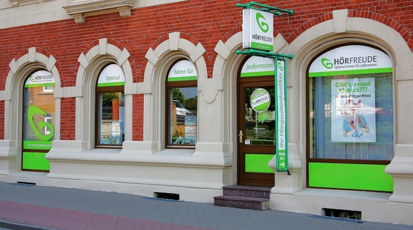 Fassade eines Hörakustikgeschäfts mit grün-weißer Beschriftung und Werbeplakaten.